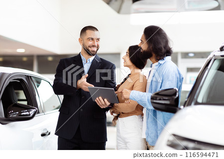A cheerful salesman, holding a digital tablet, is engaging with Indian couple next to a new car at a vehicle dealership. The man and woman appear interested and attentive 116594471