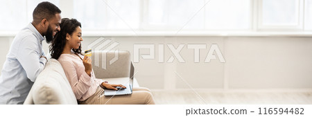 Online Shopping From Home, E-commerce Concept. Happy young african american couple sitting on sofa at home and buying clothes or products online, paying with credit card using laptop, copy space 116594482