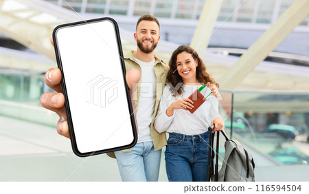 A beaming young man and woman stand inside a bustling airport terminal, showcasing a large, blank smartphone screen. 116594504