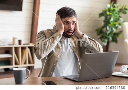 A man is seated in front of a laptop with a look of concentration, holding his head in frustration or contemplation. He appears to be deep in thought or experiencing some sort of mental strain. 116594588