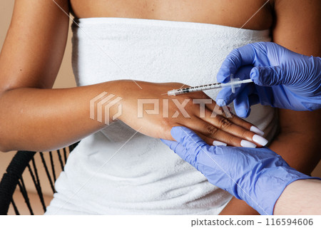 A medical professional is administering a syringe injection into the hand of a woman. The woman appears to be calmly receiving the medical procedure in a clinical setting, cropped 116594606