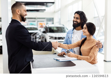 A man and a woman are shaking hands inside a well-lit car showroom. The man appears to be a car salesman, and Indian woman is a potential customer. They are standing next to a shiny car A man and a woman are shaking hands inside a well-lit car showroom. The man appears to be a car salesman, and Indian woman is a potential customer. They are standing next to a shiny car 116594613