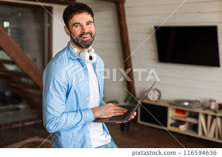 A cheerful man with a beard is standing in a well-lit home office, wearing a casual blue shirt with headphones around his neck and holding an open laptop in one hand, suggesting a break from work 116594636