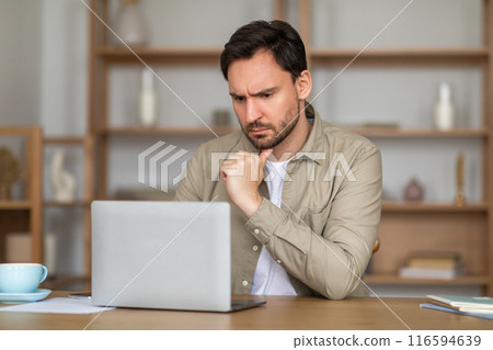 A man appears deep in thought as he works on a laptop placed on a wooden table. His body language exudes concentration and possibly some concern, as he rests his chin on his hand. 116594639