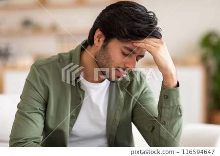 Arab man is seated on a couch, leaning back with his hand on his head. He appears deep in thought or contemplation, with a pensive expression on his face 116594647