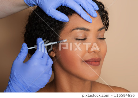 A woman is undergoing filler injection treatment on her facial skin. The injection is being administered by a trained professional to reduce the appearance of wrinkles and fine lines on her face. A woman is undergoing filler injection treatment on her facial skin. The injection is being administered by a trained professional to reduce the appearance of wrinkles and fine lines on her face. 116594656