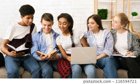 Five multiethnic teenagers are sitting closely together, sharing a casual study session indoors. Two of them are reading books, one is typing on a laptop, and the others appear to be discussing Five multiethnic teenagers are sitting closely together, sharing a casual study session indoors. Two of them are reading books, one is typing on a laptop, and the others appear to be discussing 116594721