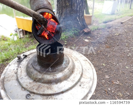 Lighting a camping samovar for making hot drinks like tea, coffee in nature near the lake by adding burning coals. Equipment for hiking, survival, long trips, tourism Lighting a camping samovar for making hot drinks like tea, coffee in nature near the lake by adding burning coals. Equipment for hiking, survival, long trips, tourism 116594739