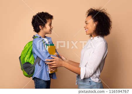 A young African American boy, outfitted with a green backpack and holding colorful books, stands facing his smiling mother as she offers him support and affirmation before he heads off to school A young African American boy, outfitted with a green backpack and holding colorful books, stands facing his smiling mother as she offers him support and affirmation before he heads off to school 116594767