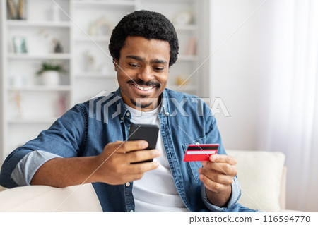African American man is seated on a couch, holding a smart phone in one hand and a credit card in the other, focused on the phone screen as he possibly makes an online purchase 116594770