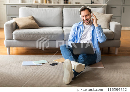 A man is sitting on the floor, wearing headphones and attentively listening to music while using a laptop. He appears focused and engaged in his digital activities. 116594832