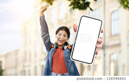 Hindu woman is standing and holding up a smartphone with a blank screen. She is looking at the phone intently, possibly waiting for a response or checking for notifications. 116594842