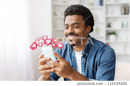 Black man with a joyful expression smiles as he looks at his smartphone. He is clearly pleased with the notifications he is receiving, which are represented by heart-shaped icons floating above Black man with a joyful expression smiles as he looks at his smartphone. He is clearly pleased with the notifications he is receiving, which are represented by heart-shaped icons floating above 116594880