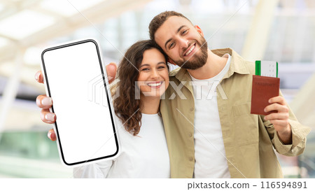 A cheerful man and woman are shown close together holding up a smartphone with a blank screen and a passport. They seem to be in a well-lit airport, possibly ready for travel A cheerful man and woman are shown close together holding up a smartphone with a blank screen and a passport. They seem to be in a well-lit airport, possibly ready for travel 116594891