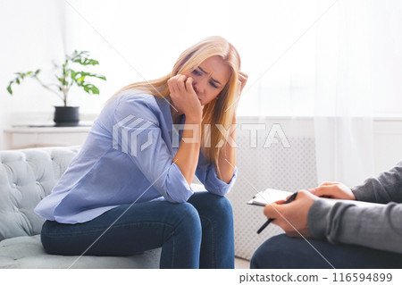 A woman sits on the edge of a couch, her hand covering her face, possibly wiping tears, expressing distress or frustration during a counseling session A woman sits on the edge of a couch, her hand covering her face, possibly wiping tears, expressing distress or frustration during a counseling session 116594899