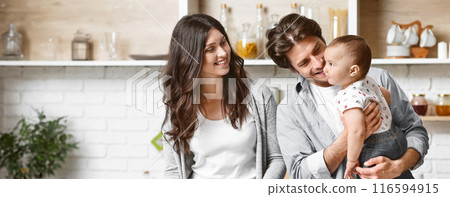 A young family smiles in their modern kitchen. The father holds their baby while the mother looks on with a loving expression. The kitchen features white brick walls and open shelves. 116594915