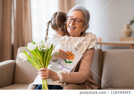 A young girl with braided hair is hugging her glasses-wearing grandmother, who is sitting on a beige sofa. The child is presenting a bouquet of white tulips and a handmade card 116594945