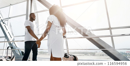 African American couple walks hand-in-hand through an airport terminal, luggage in tow. They appear to be heading towards their gate, excited for their journey, copy space 116594956