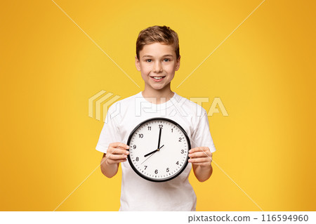 A young boy holds a clock in front of his face, showcasing the time with a serious expression. 116594960