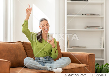 A woman sits cross-legged on a tan leather couch, with a triumphant expression and her arm raised in a victory gesture. She holds a smartphone in her other hand, possibly having received good news A woman sits cross-legged on a tan leather couch, with a triumphant expression and her arm raised in a victory gesture. She holds a smartphone in her other hand, possibly having received good news 116595001