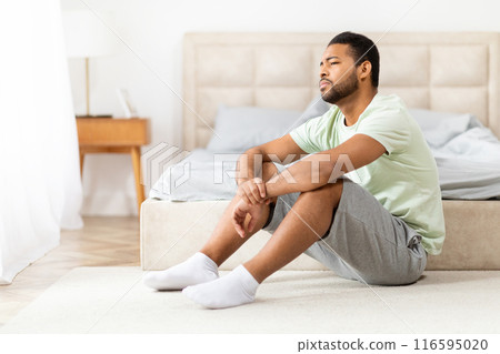 A young African American man sits on the floor next to bed in a sunlit bedroom, he looks upset, frustrated, depressed, copy space 116595020