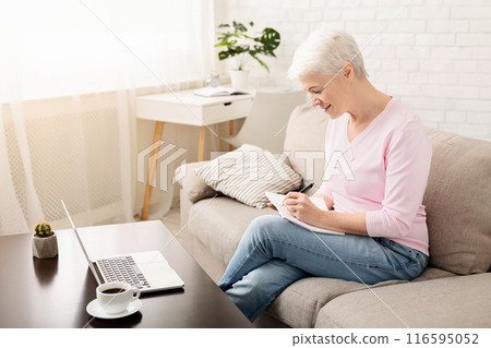 A smiling senior woman with short grey hair is sitting comfortably on a beige couch in a bright living room, engaging with her laptop, taking notes 116595052