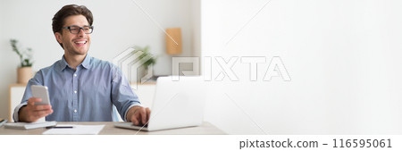 happy young businessman wearing eyeglasses using phone and laptop while working at home office, panorama with copy space 116595061