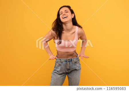 A cheerful young woman wearing a pastel pink tank top and blue denim jeans stands against a vivid yellow backdrop, her hands on her hips as she tilts her head back in a spontaneous laugh A cheerful young woman wearing a pastel pink tank top and blue denim jeans stands against a vivid yellow backdrop, her hands on her hips as she tilts her head back in a spontaneous laugh 116595083