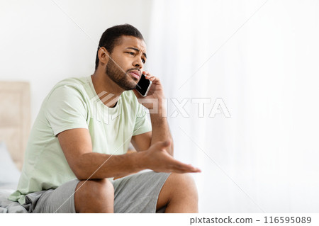A young African American man is seated on his bed with a serious expression, talking on his cellphone. The room appears bright and minimalistic, copy space A young African American man is seated on his bed with a serious expression, talking on his cellphone. The room appears bright and minimalistic, copy space 116595089