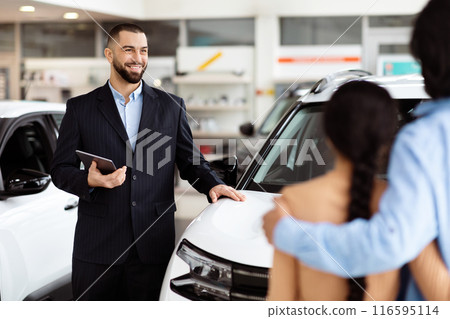 A professional salesman, clad in a smart suit, stands by a shiny new car with a digital tablet in hand, confidently explaining features to an interested Indian couple in a car dealership showroom A professional salesman, clad in a smart suit, stands by a shiny new car with a digital tablet in hand, confidently explaining features to an interested Indian couple in a car dealership showroom 116595114