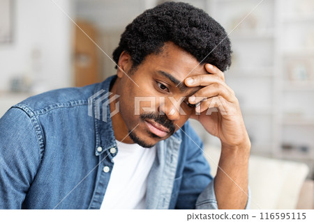 Upset African American man is seated on a couch, with his hand resting on his head in a thoughtful gesture. He appears deep in contemplation or possibly experiencing stress or fatigue Upset African American man is seated on a couch, with his hand resting on his head in a thoughtful gesture. He appears deep in contemplation or possibly experiencing stress or fatigue 116595115