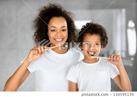 African American mother and her young child, both donning white t-shirts, share a cheerful moment while brushing their teeth together in a well-lit bathroom 116595130