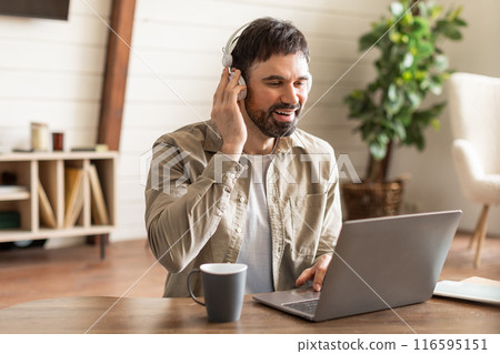 A man is seated at a table, focused on his laptop screen while wearing headphones. He appears to be working or studying intently. 116595151