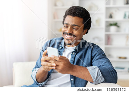 African American man is seated on a couch, engrossed in his cell phone, scrolling through content displayed on the screen. 116595152