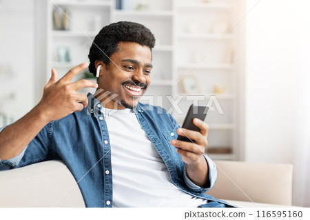 A cheerful African American man is engaging in a video call, making a hand gesture as if he speaking to someone while relaxing on a comfortable sofa A cheerful African American man is engaging in a video call, making a hand gesture as if he speaking to someone while relaxing on a comfortable sofa 116595160