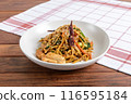 A savory plate of stir-fried noodles garnished with sesame seeds and chili peppers, served on a wooden table with a checkered napkin 116595184
