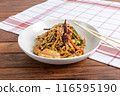 A plate of stir-fried noodles mixed with chicken, vegetables, and topped with sesame seeds, served on a wooden table with chopsticks 116595190