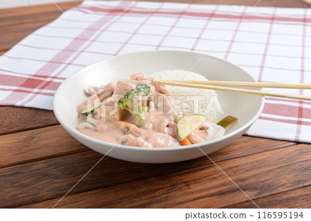 A white plate with Thai chicken curry and jasmine rice served on a wooden table with chopsticks resting on a checkered napkin 116595194