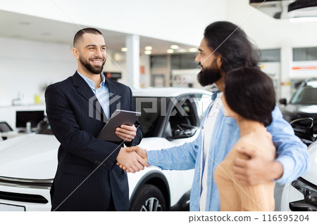 A well-dressed salesman is shaking hands with happy Indian customers, presumably after agreeing on a sale, with a new white car in the background inside a modern car dealership showroom. 116595204