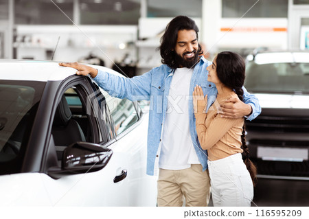Indian man and woman standing next to a parked car. The man is wearing a blue shirt and jeans, while the woman is in a red dress. They appear to be discussing something, with the car door open. 116595209