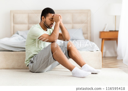 A young African American man with a look of discomfort or stress is seated on the floor by bed in a well-lit, modern bedroom, copy space 116595418