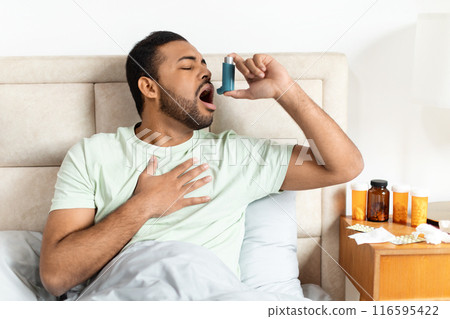 African American young man is sitting on the edge of his bed, holding his chest with one hand while administering medication with an asthma inhaler into his open mouth 116595422