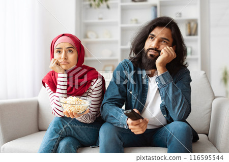 Unhappy indian young couple is lounging on a cozy sofa in a well-lit living room, seemingly unimpressed or bored while holding a bowl of popcorn. 116595544
