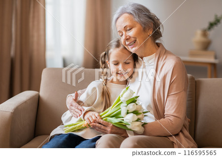 A tender moment is captured as a smiling grandmother wraps her arms around her young granddaughter, who is holding a bouquet of fresh tulips A tender moment is captured as a smiling grandmother wraps her arms around her young granddaughter, who is holding a bouquet of fresh tulips 116595565