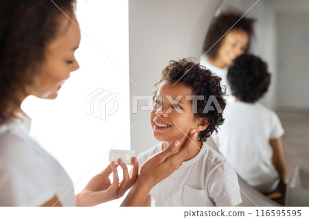 African American mother tenderly applies sunscreen to her child cheerful face, ensuring protection from the suns rays. Both are dressed in casual white attire, reflecting a carefree, sunny day. African American mother tenderly applies sunscreen to her child cheerful face, ensuring protection from the suns rays. Both are dressed in casual white attire, reflecting a carefree, sunny day. 116595595