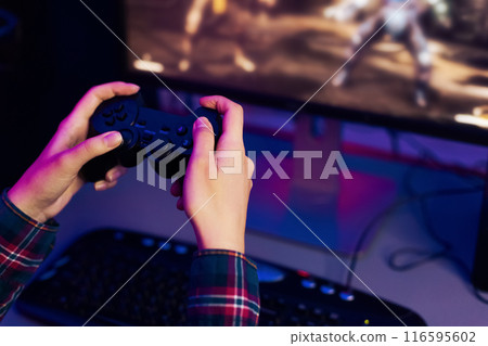 This is a photo of a persons hands holding a video game controller, with a computer monitor in the background displaying an action game. The person is immersed in the game, focused on the screen. This is a photo of a persons hands holding a video game controller, with a computer monitor in the background displaying an action game. The person is immersed in the game, focused on the screen. 116595602