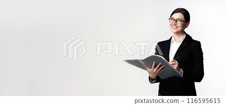 A businesswoman in a black suit and glasses smiles as she writes in a notebook. She stands against a white background. A businesswoman in a black suit and glasses smiles as she writes in a notebook. She stands against a white background. 116595615
