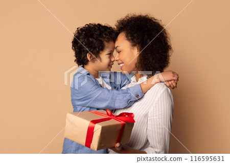 Cute African American little boy greeting his mom with Mothers Day, giving her wrapped box and embracing his mummy, beige studio background 116595631