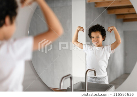 African American young child is standing in front of a bathroom mirror, proudly flexing their muscles and exhibiting a joyful smile. The bathroom has a modern design with visible wooden beams 116595636
