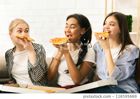 Three young girls are seated on a couch, enjoying slices of pizza. The girls are casually dressed and appear to be having a relaxed and enjoyable time together. 116595669